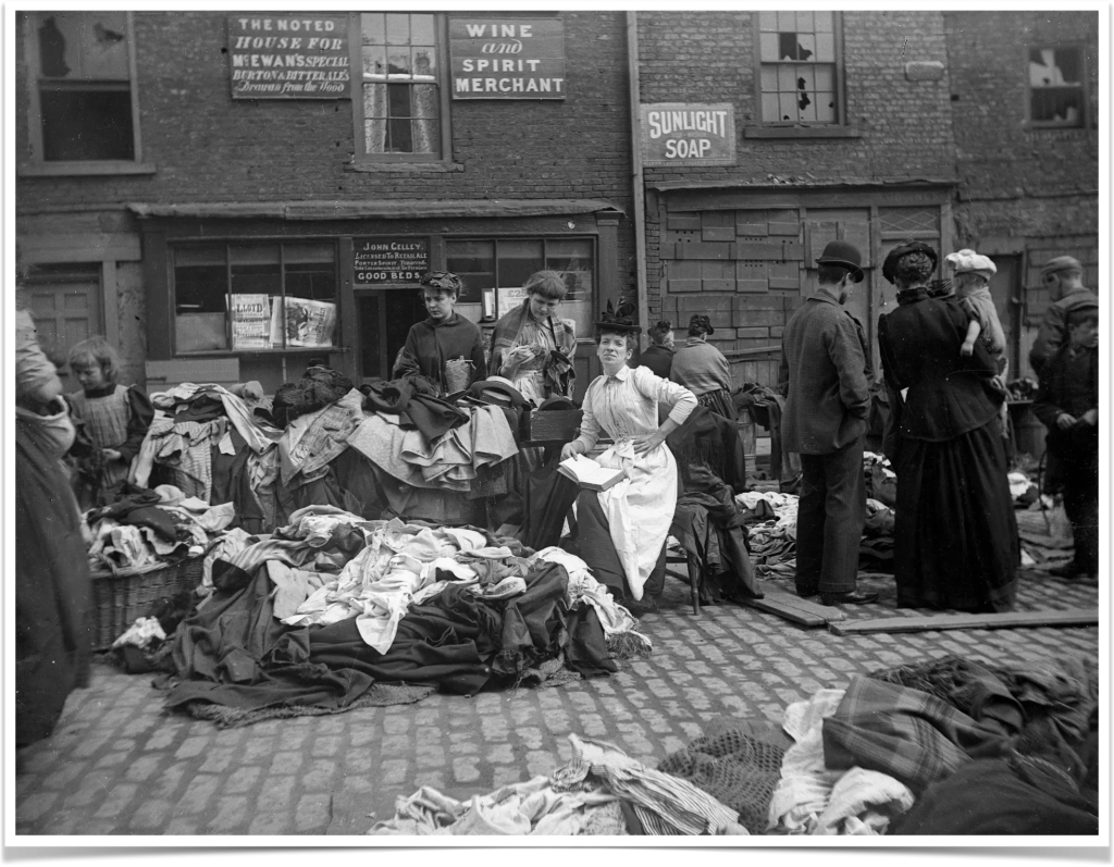 A secondhand clothes seller at Newcastle Cloth Market reads as customers inspect her goods, mid 19th century. Photograph: NEMiPA- Collection of the Society of Antiquaries, Newcastle